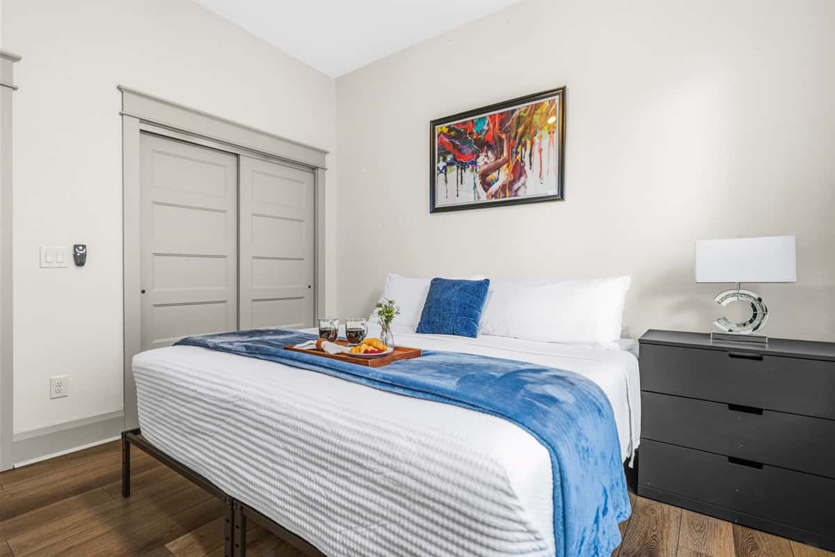 Stylish bedroom with a navy blue accent comforter, built-in closet, dark wood dresser, vibrant wall art, and a wooden breakfast tray. Brand-new finishes and hardwood floors make every bedroom feel like a retreat.