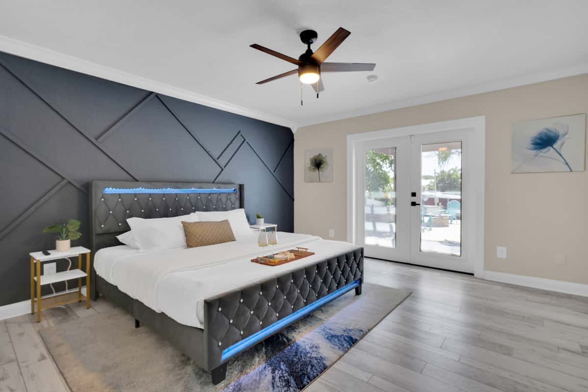 The primary suite's tufted bed and moody accent wall open straight through French doors to the pool patio, indoor comfort meets outdoor paradise