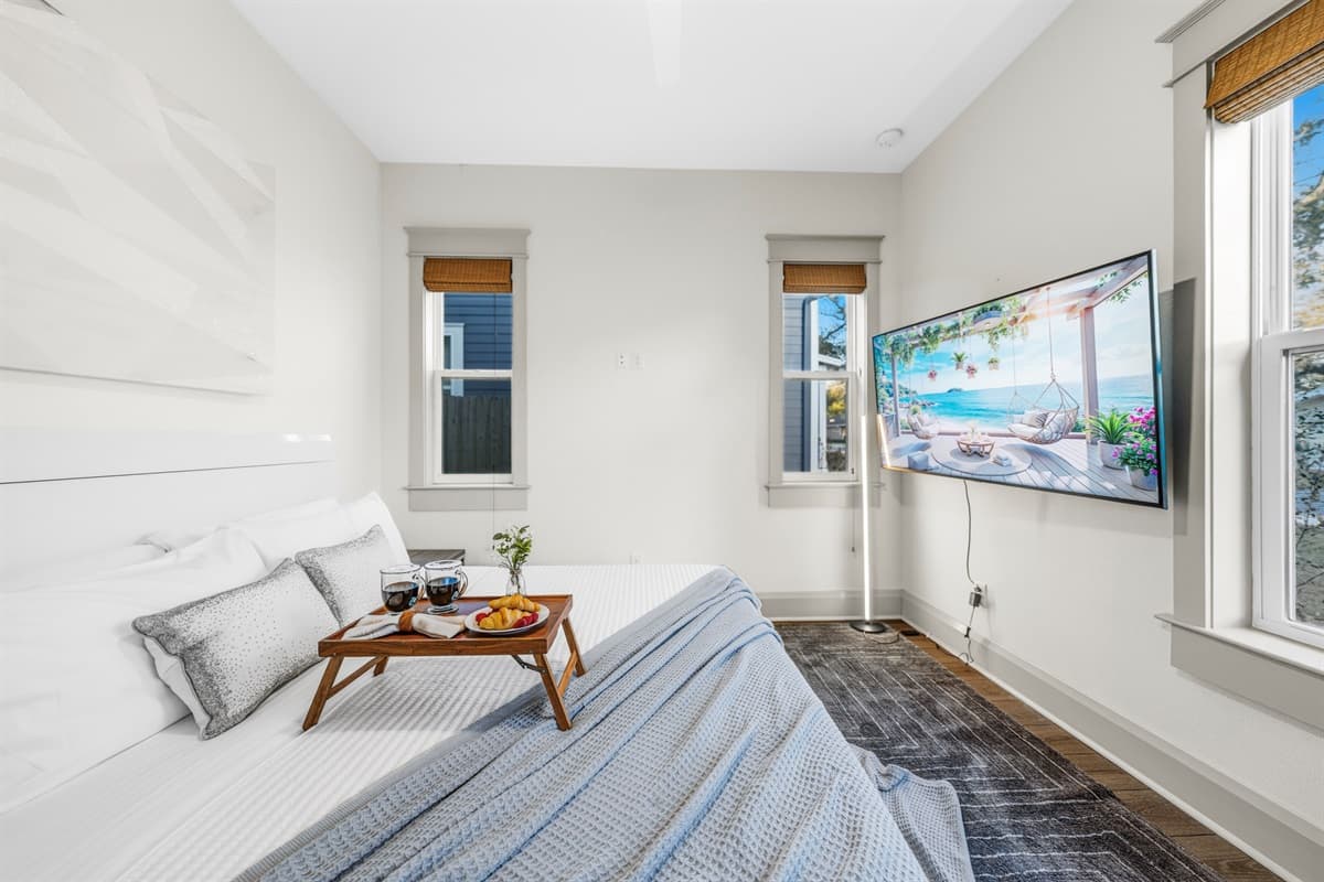 Cozy bedroom with a wall-mounted smart TV, wooden breakfast tray, soft blue comforter, and a plush area rug over hardwood floors. Everything you need to relax and recharge in this brand-new Seminole Heights retreat.