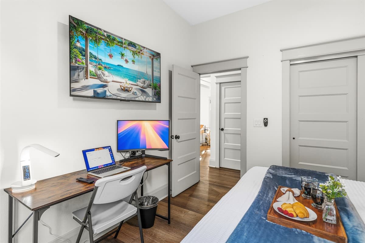 Another angle of this gorgeous bedroom — tropical wall art, a dedicated desk workspace, blue accent bedding, breakfast tray with fresh fruit, and gleaming hardwood floors. Brand-new and ready for your best stay yet.