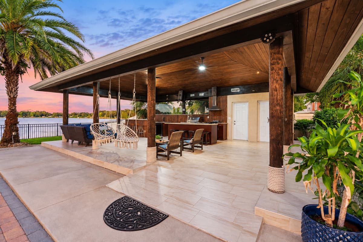 A spacious covered patio area overlooking the water at sunset. Surrounded by palm trees and tropical greenery, the patio offers a relaxing waterfront setting perfect for outdoor dining and gathering with family and friends.