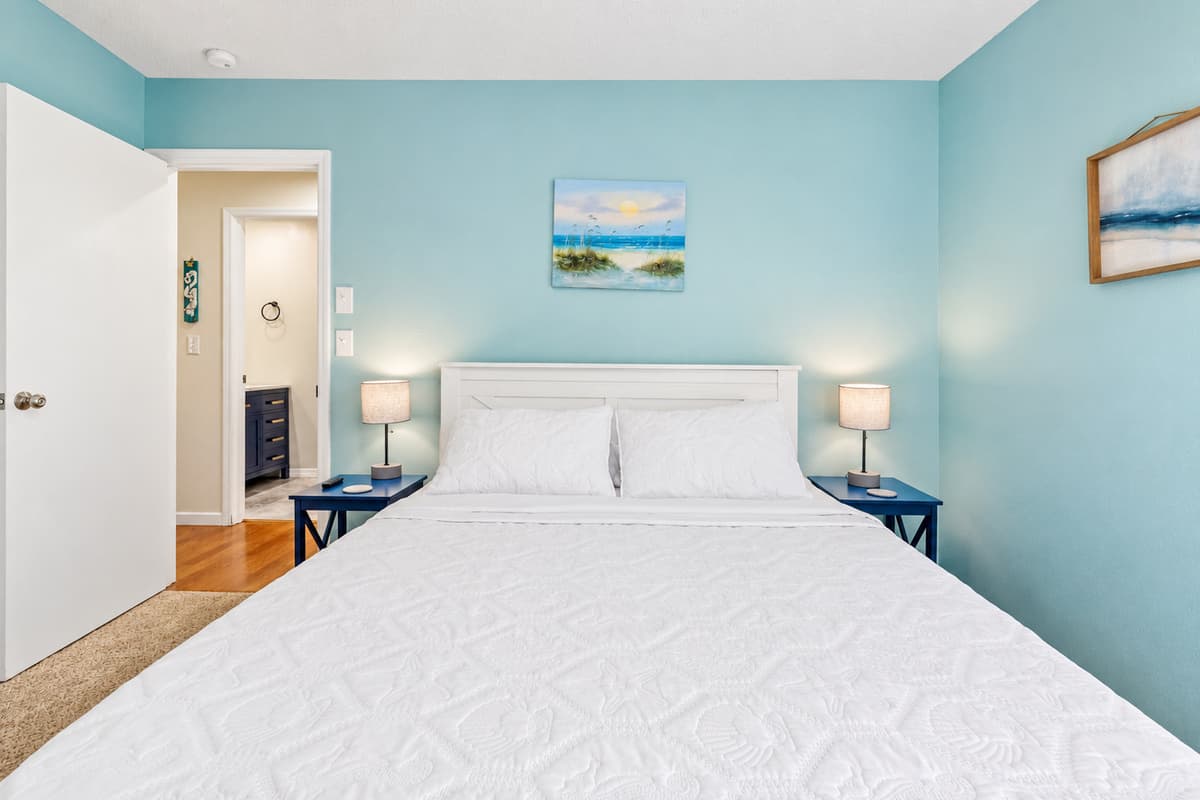 The walls are painted a cool, serene coral blue, evoking the calm and refreshing vibes of the nearby ocean, and are perfectly complemented by the fresh, crisp white linens that dress the bed. 