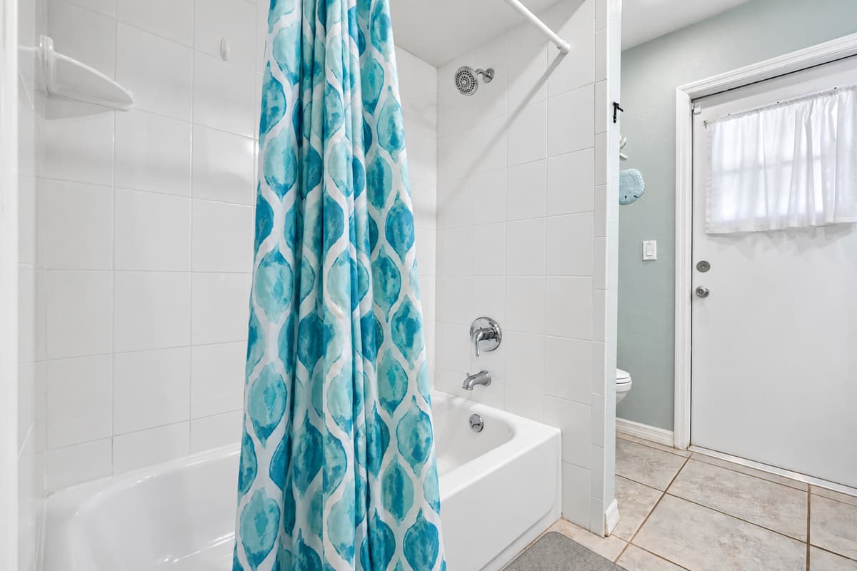 This full bathroom features a relaxing coastal design with dual vanities, granite countertops, and a beautifully tiled shower/tub combo accented by a vibrant blue curtain.