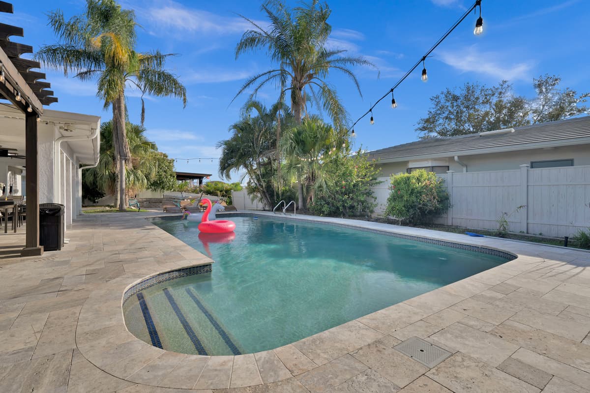 Sunlit pool with a floating flamingo and travertine pavers underfoot, the kind of scene that sells the vacation before guests even arrive