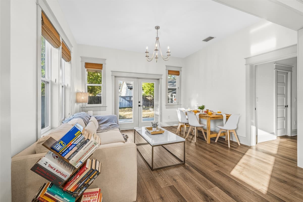 Sun-drenched sitting room bathed in natural light, featuring a stunning chandelier, sofa with coffee table, wood blinds, and an open view to the dining area — an elegant and relaxing space in this brand-new home.