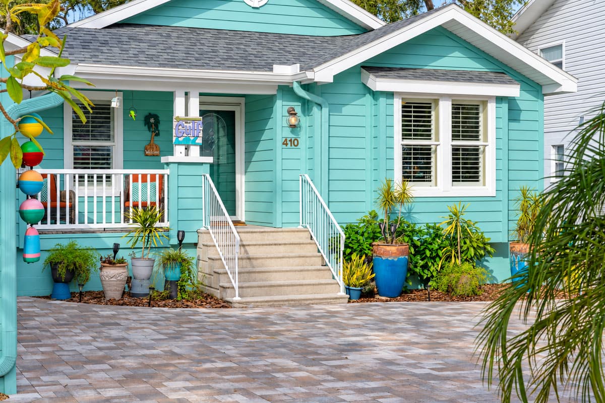 This bright and airy home showcases a stunning paver driveway, charming porch seating, and tropical touches throughout.