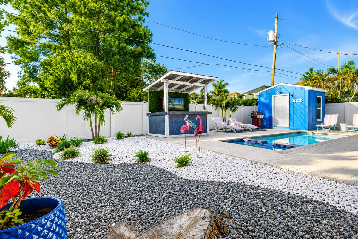 Wide view of the private backyard oasis showcasing the pool, lounge area, outdoor bar, tropical landscaping, and colorful coastal accents.