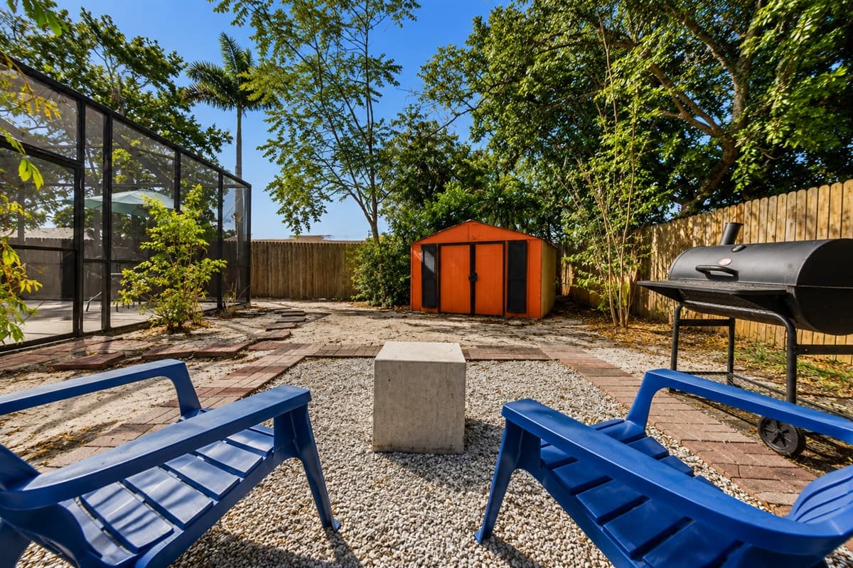 Scenic backyard view showcasing the fire pit seating area, grill station, and surrounding tropical landscaping.