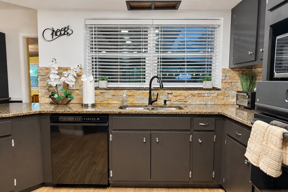 This sleek, dark-toned kitchen makes a stylish statement with black cabinetry, warm stone backsplash, and wood-look floors—perfect for whipping up a feast or morning coffee.