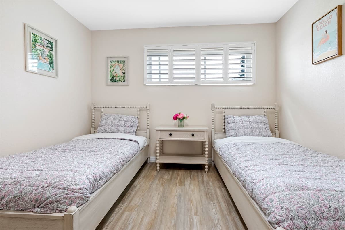 This cozy bedroom features two matching twin beds with light wood frames, a central nightstand with fresh flowers, and cheerful wall art that adds a pop of color.