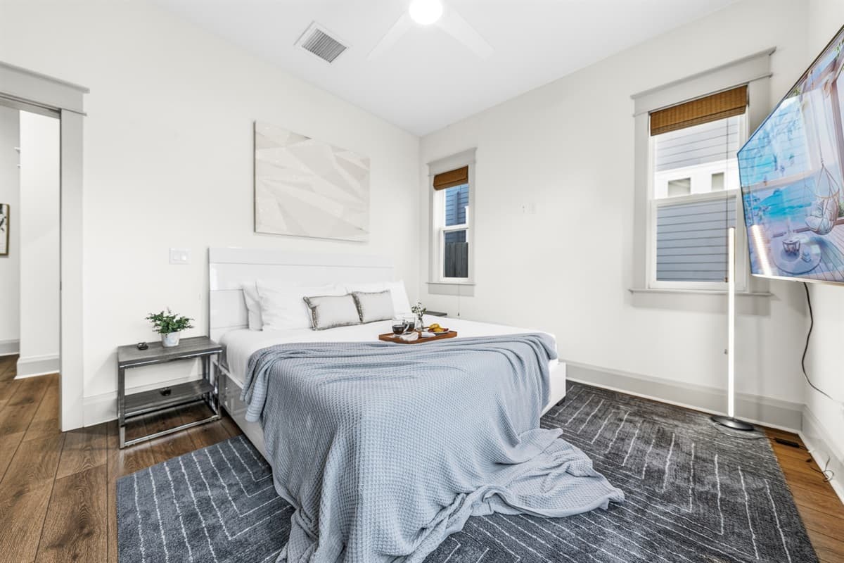 Serene and spacious bedroom with a king bed, soft gray linen, ceiling fan, plush area rug, nightstand with fresh greenery, and a bamboo-shaded window. Brand-new tranquility in the heart of Seminole Heights.