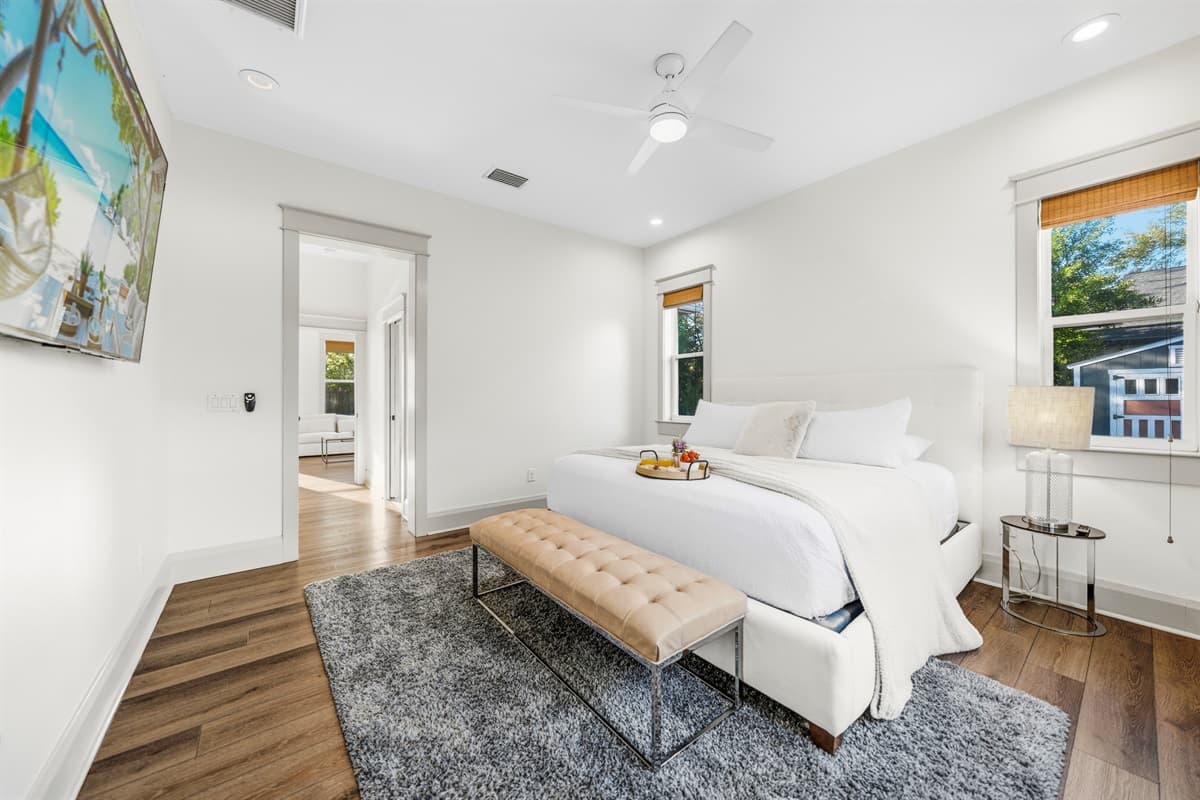 Spacious bedroom with a bold tropical beach canvas, tufted bench, gray area rug, hardwood floors, and soaring ceilings. This brand-new Seminole Heights home brings vacation vibes to every room.