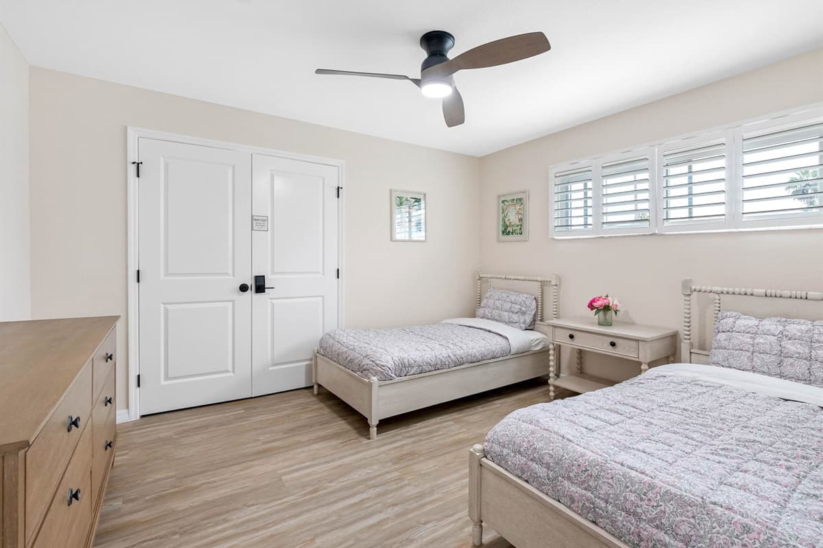 A simple, charming space with twin beds, bright windows with shutters, and a touch of greenery in framed art for a refreshing atmosphere.
