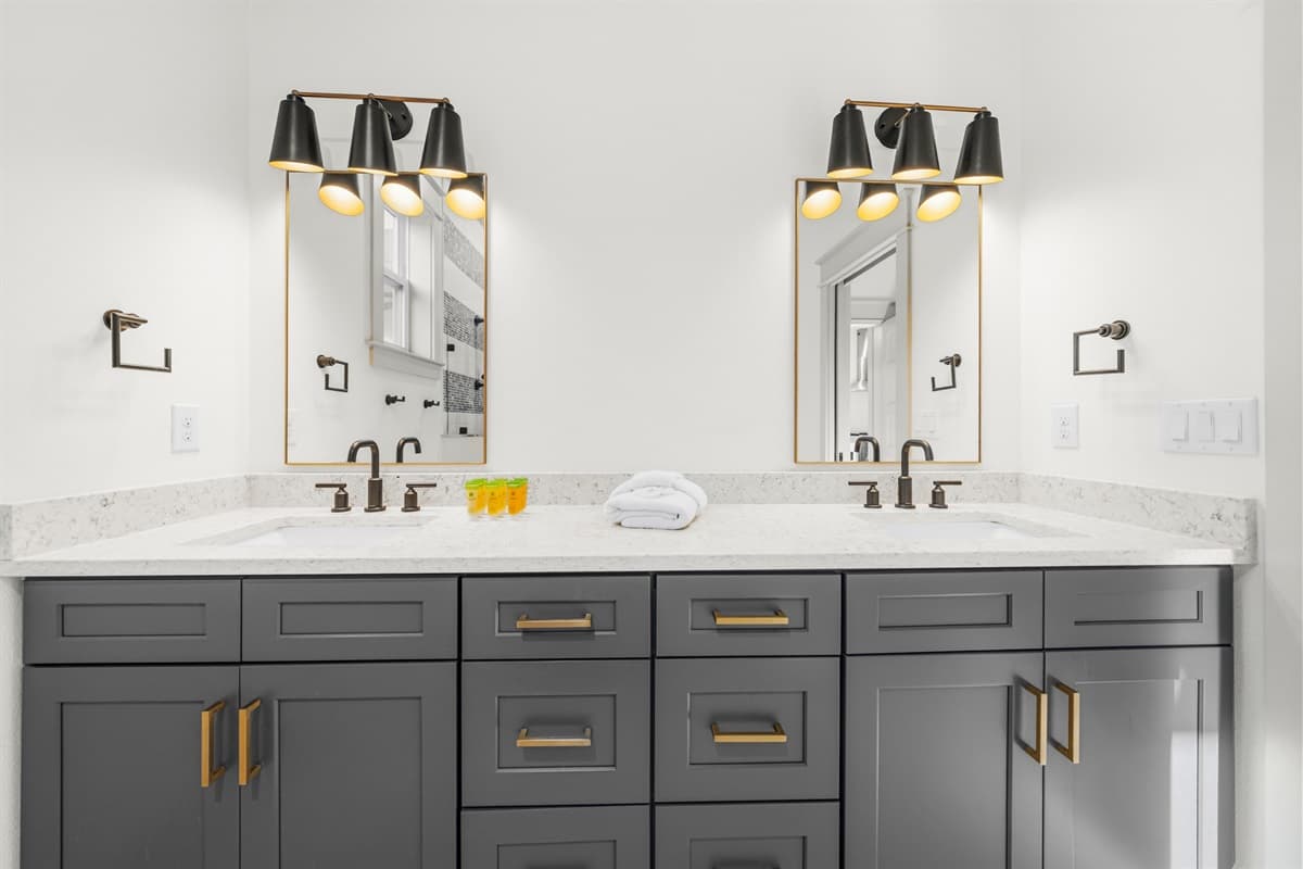 Stunning double vanity bathroom with dark charcoal cabinets, gold hardware, double white quartz sinks, backlit mirror, and elegant black and gold sconce lighting. Brand-new luxury in the heart of Seminole Heights.