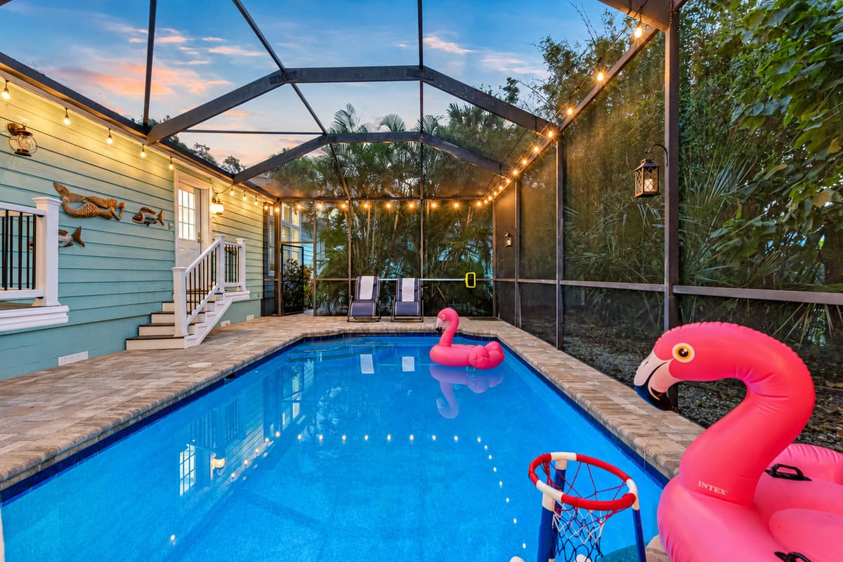 Enjoy a tropical atmosphere with glowing string lights, vibrant pool floats, and lush vegetation bordering the screened pool.