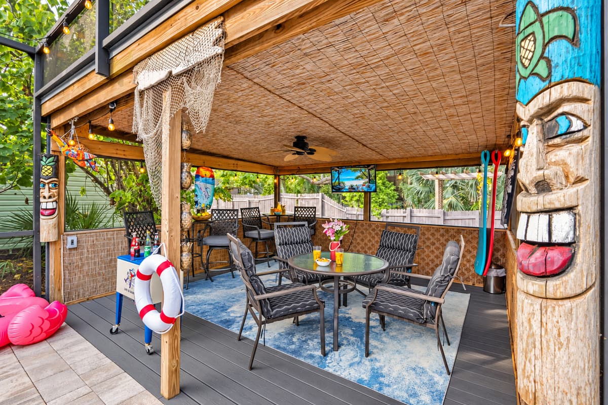 A tropical-themed covered patio featuring tiki décor, string lights, and multiple seating areas beside the pool.