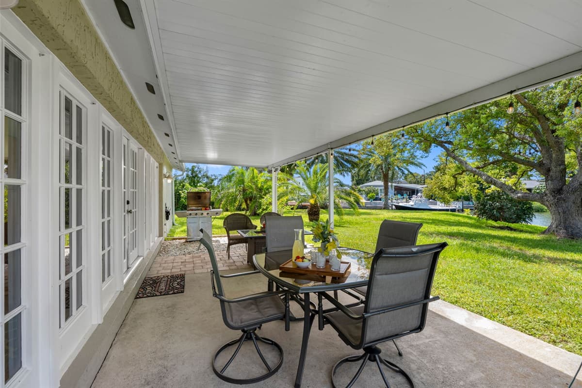 This patio blends indoor comfort with outdoor beauty, featuring a shaded roof, stylish furniture, and a scenic water view.
