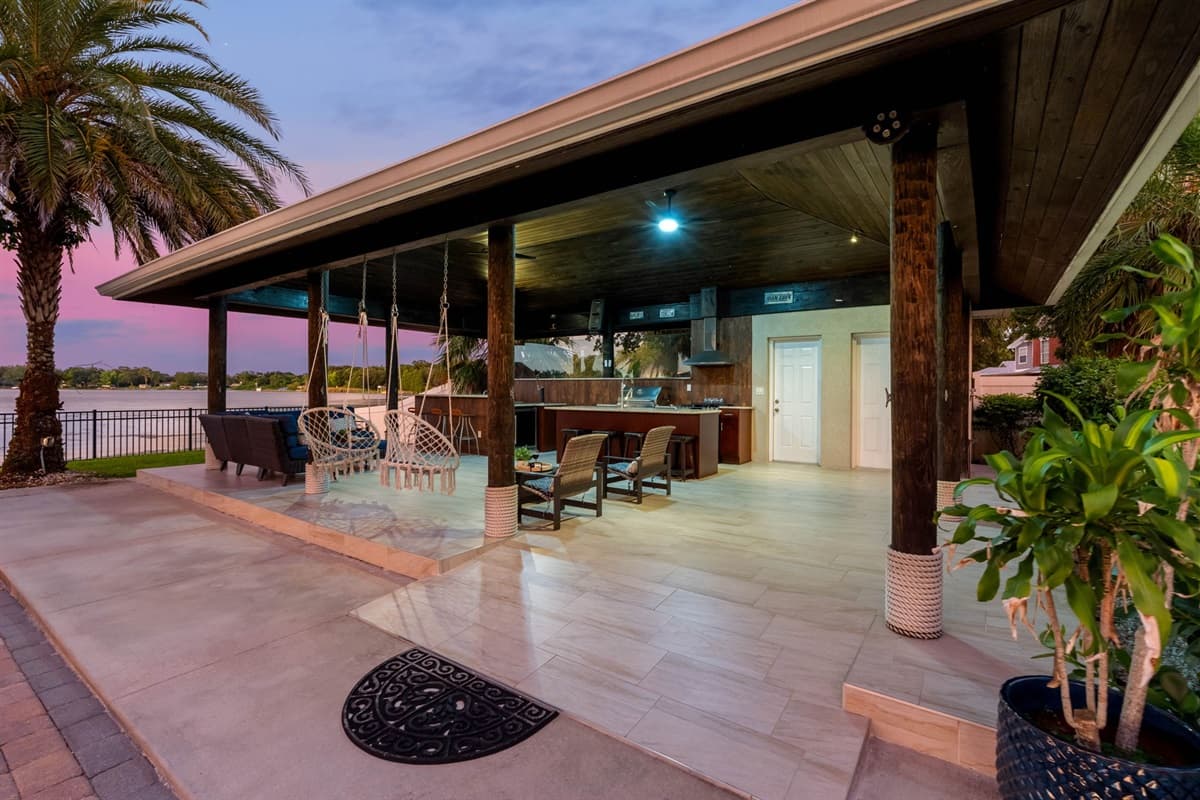 A spacious covered patio area overlooking the water at sunset. Surrounded by palm trees and tropical greenery, the patio offers a relaxing waterfront setting perfect for outdoor dining and gathering with family and friends.