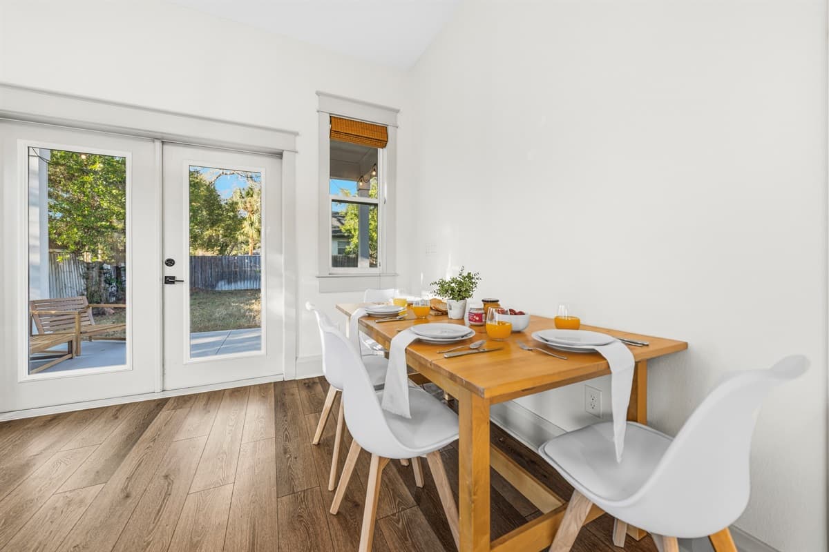Warm and welcoming dining space with a solid wood farmhouse table, sleek white modern chairs, and floor-to-ceiling French doors overlooking the lush greenery of Seminole Heights. Meals here feel like a special occasion every time.