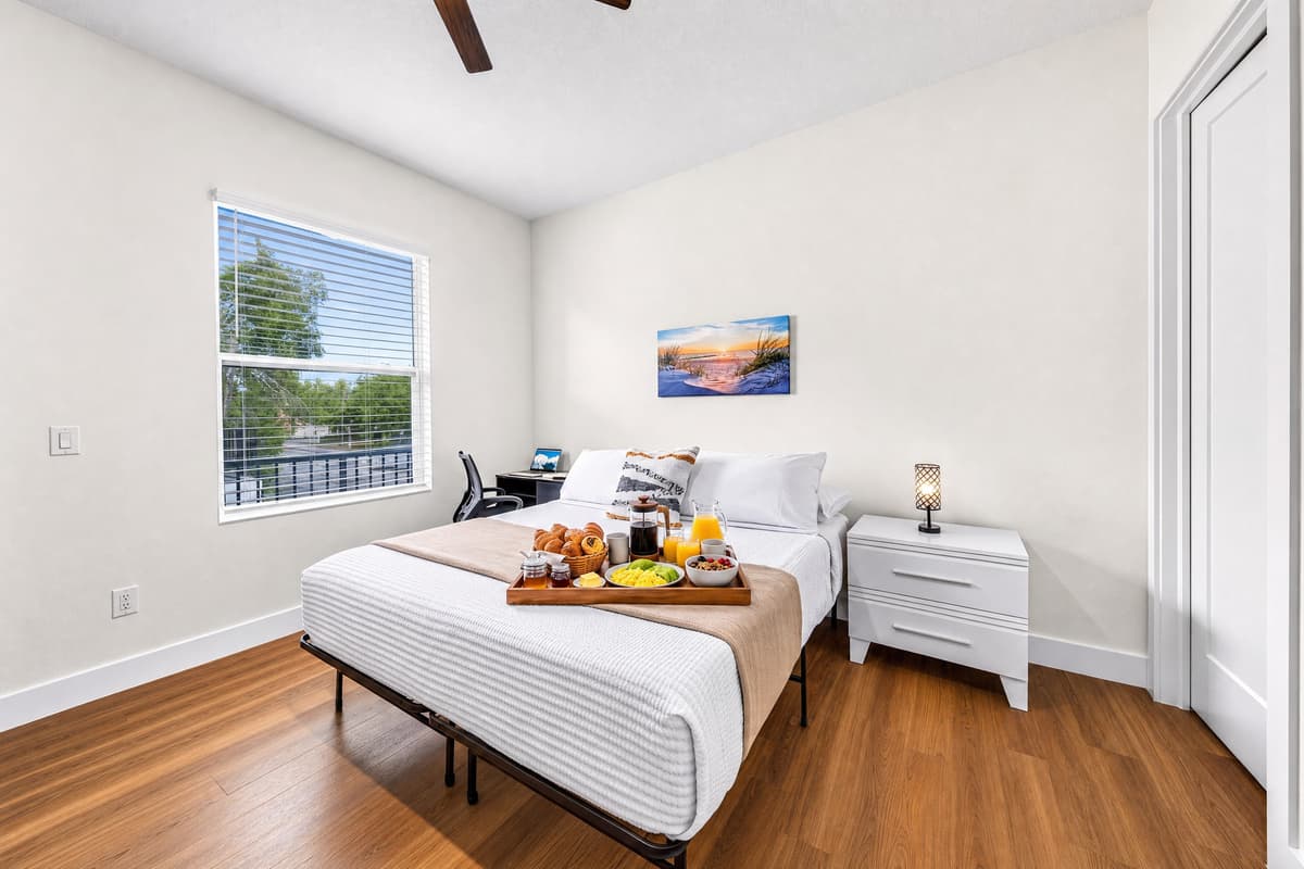 Your serene retreat: a queen bed, work desk, beach sunset art, and treetop views through the window. Work or relax — your choice.
