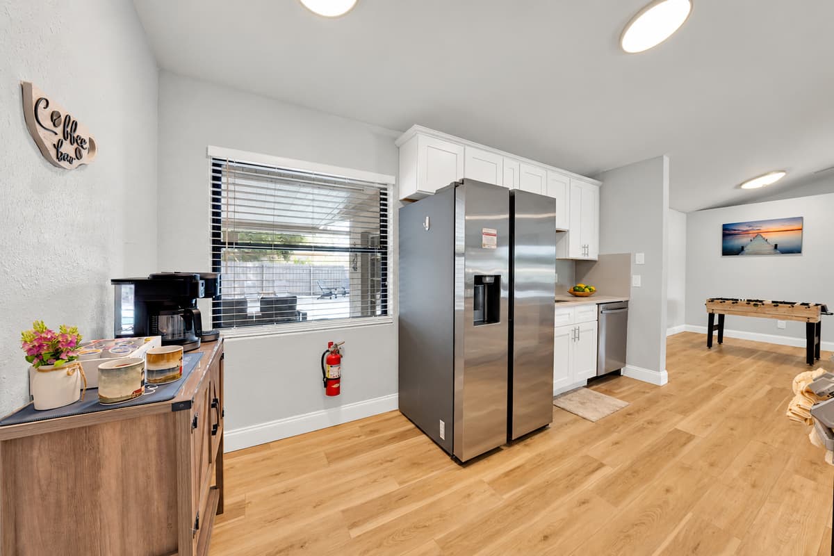 Coffee bar on one side, full-size stainless fridge with ice maker on the other, and the dining table already in view down the hall. Everything the morning needs sits within about six steps of itself.