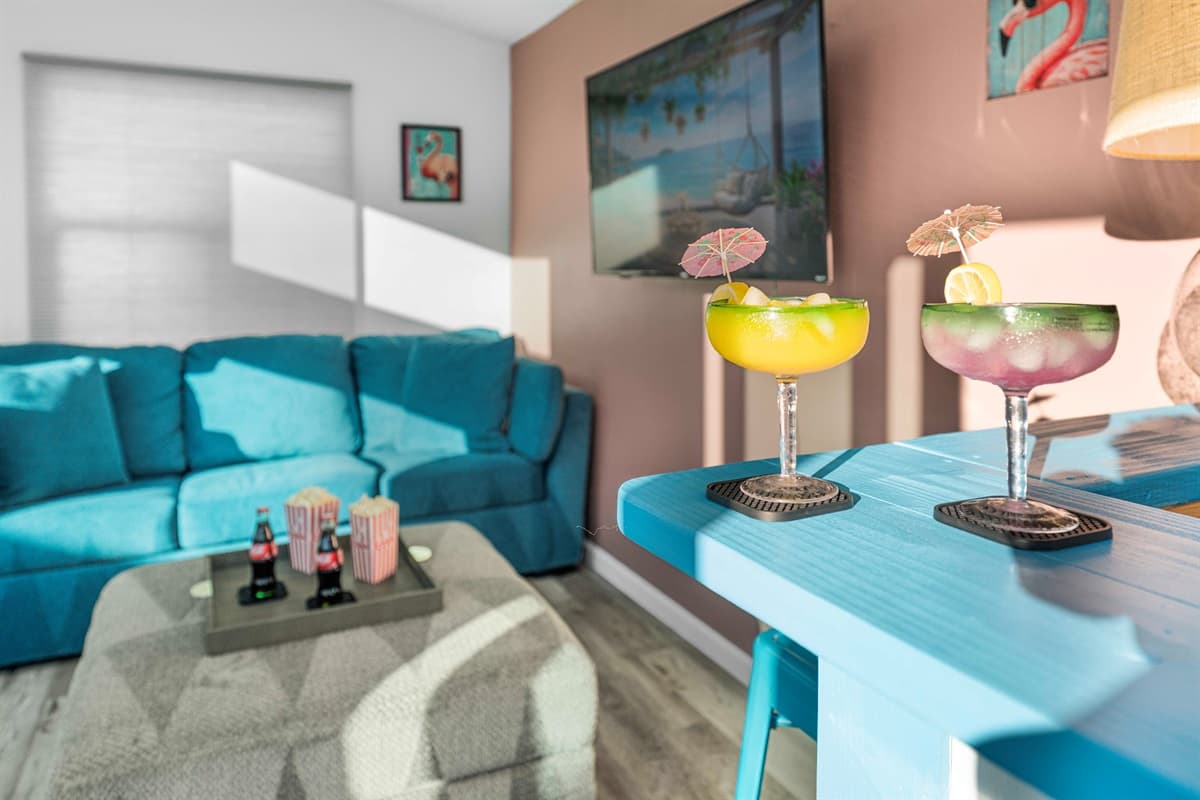 Flamingo Lounge - Featuring a plush teal sofa, soft natural lighting, and beach-inspired décor. In the foreground, two colorful tropical cocktails sit on a turquoise high-top table, adding a fun, vacation-ready vibe to the room.