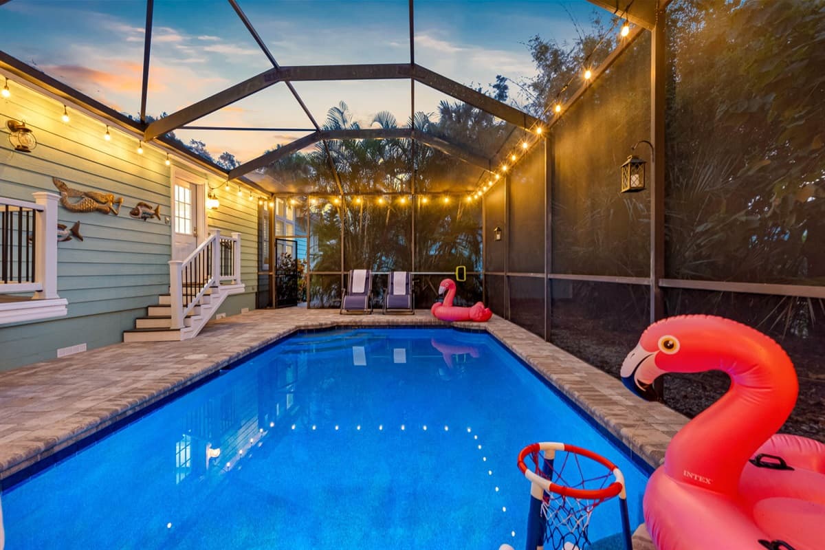 Enjoy a tropical atmosphere with glowing string lights, vibrant pool floats, and lush vegetation bordering the screened pool.