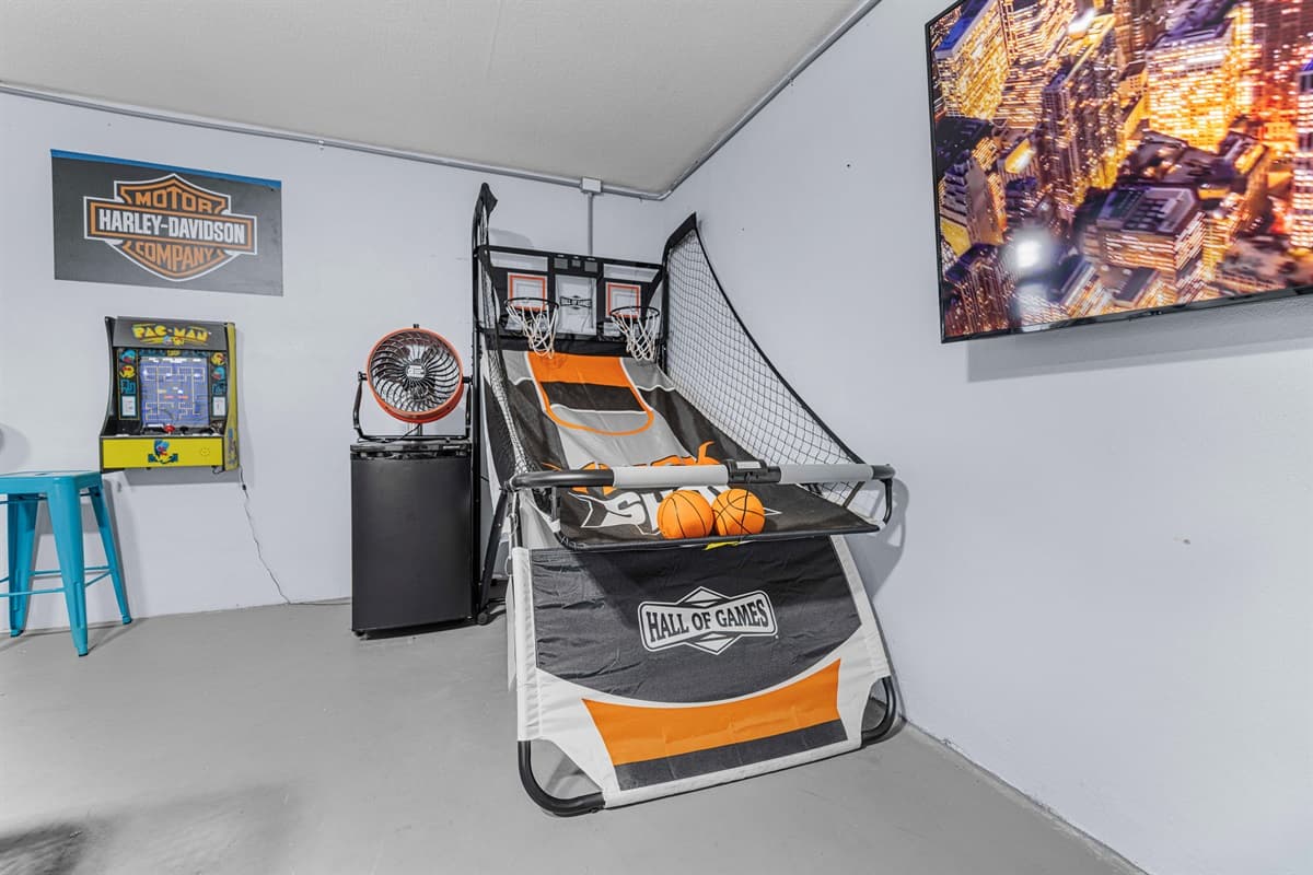 A fun-filled game room setup featuring an arcade-style basketball shooting game, a classic Pac-Man arcade machine, and a Harley-Davidson wall sign for added character. A wall-mounted TV displays a vibrant cityscape, creating an energetic atmosphere.