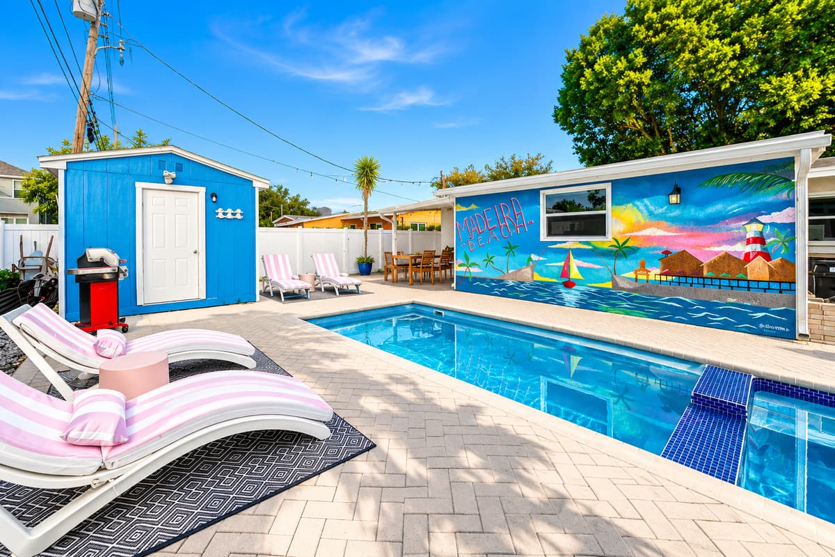 Resort-style pool area with sun loungers, paver deck, poolside shed, and vibrant beach-themed mural artwork.