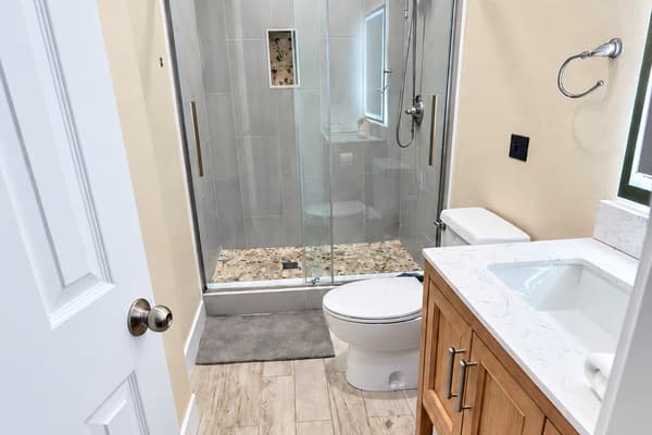 This beautifully updated bathroom features a sleek glass-enclosed shower with pebble tile accents, a modern wood vanity with a quartz countertop, and elegant brushed nickel fixtures.