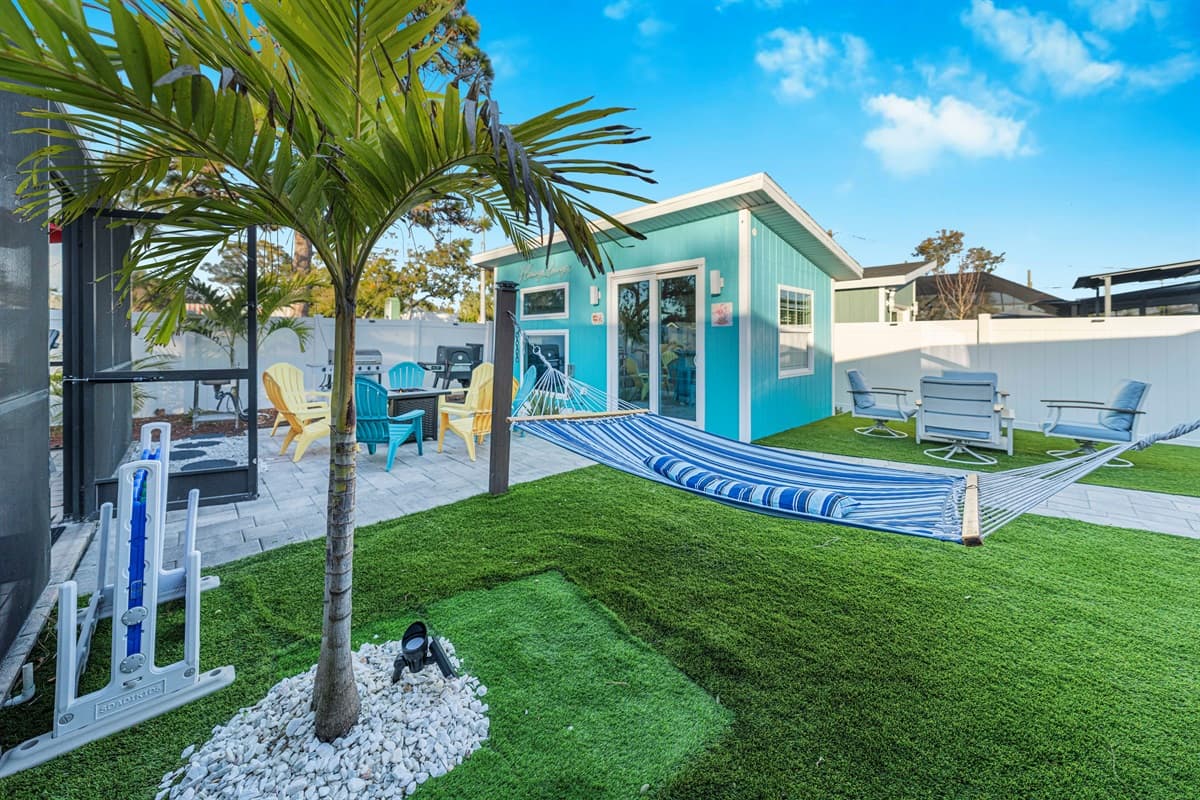 A vibrant backyard retreat with a striped hammock stretched across a beautifully maintained lawn. The turquoise exterior of the home, palm trees, and cozy patio seating create a relaxing, tropical atmosphere perfect for lounging outdoors.