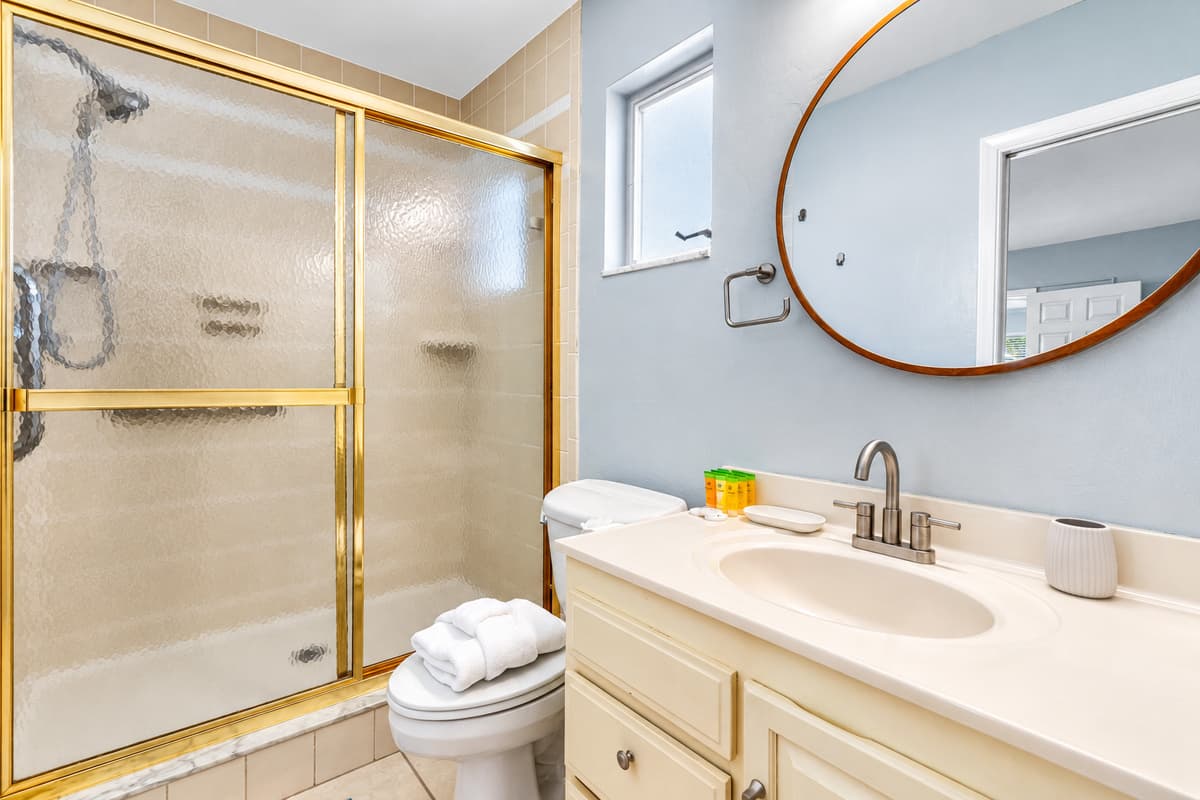 This clean and cozy bathroom features a spacious walk-in shower with gold trim, a round mirror, and natural light from the frosted window.