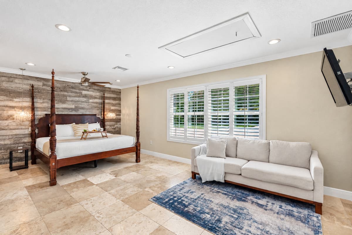 This inviting master bedroom features a grand four-poster bed, a rustic wood accent wall, and ample natural light streaming through plantation shutters.
