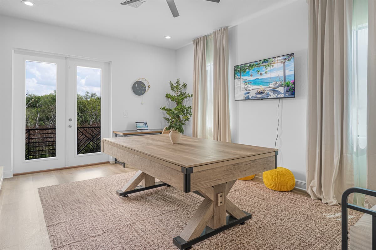 Bright entertainment room with a convertible pool table, wall-mounted TV, and direct access to outdoor views through glass doors.