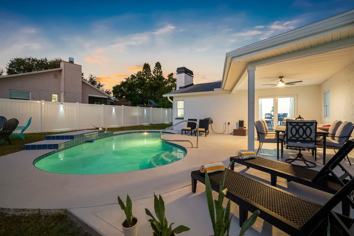 Unwind by the glowing pool as the sun sets — this private backyard retreat features comfortable loungers, outdoor dining, and serene lighting for the perfect evening ambiance.