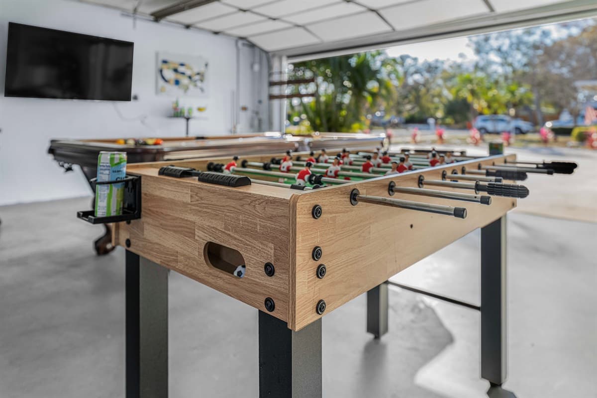 A stylish foosball table positioned in a spacious garage area with a wall-mounted TV nearby for added entertainment. The open garage door allows natural light to flow in and offers a view of the driveway and surrounding greenery.