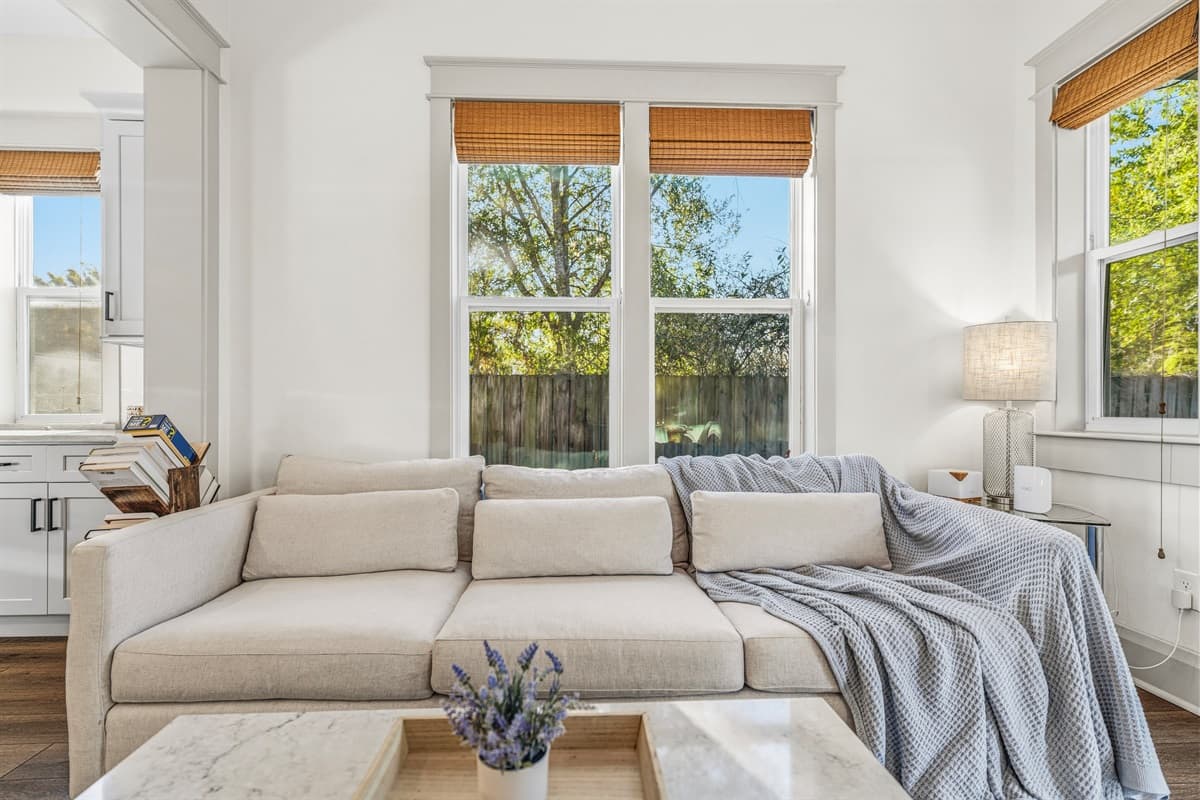 Cozy flex sitting room with a plush cream sofa, soft throw blanket, and oversized windows framing Seminole Heights' lush oak tree canopy. A peaceful retreat within this brand-new Tampa home — perfect for reading or relaxing.