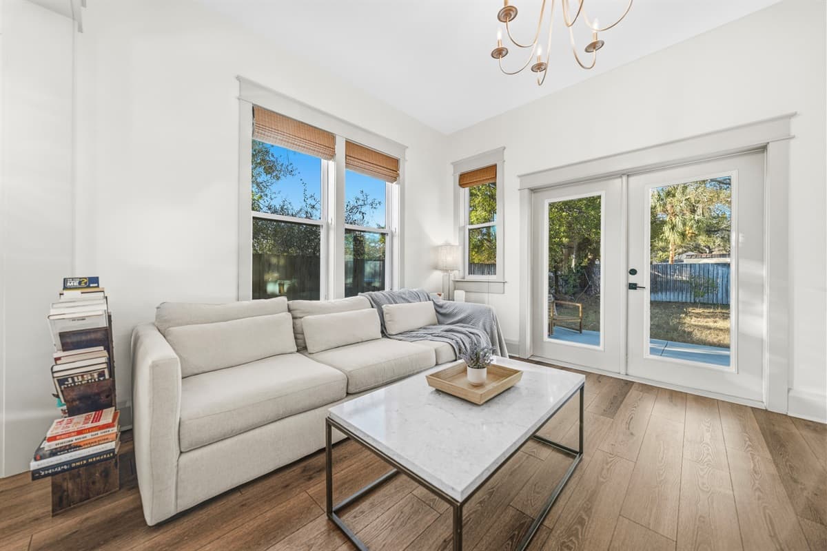 Light-filled sitting room with a gold chandelier, cream sofa, and wide French doors opening to the lush backyard. Indoor-outdoor living at its best in this brand-new Seminole Heights home — just open the doors and let Florida in.