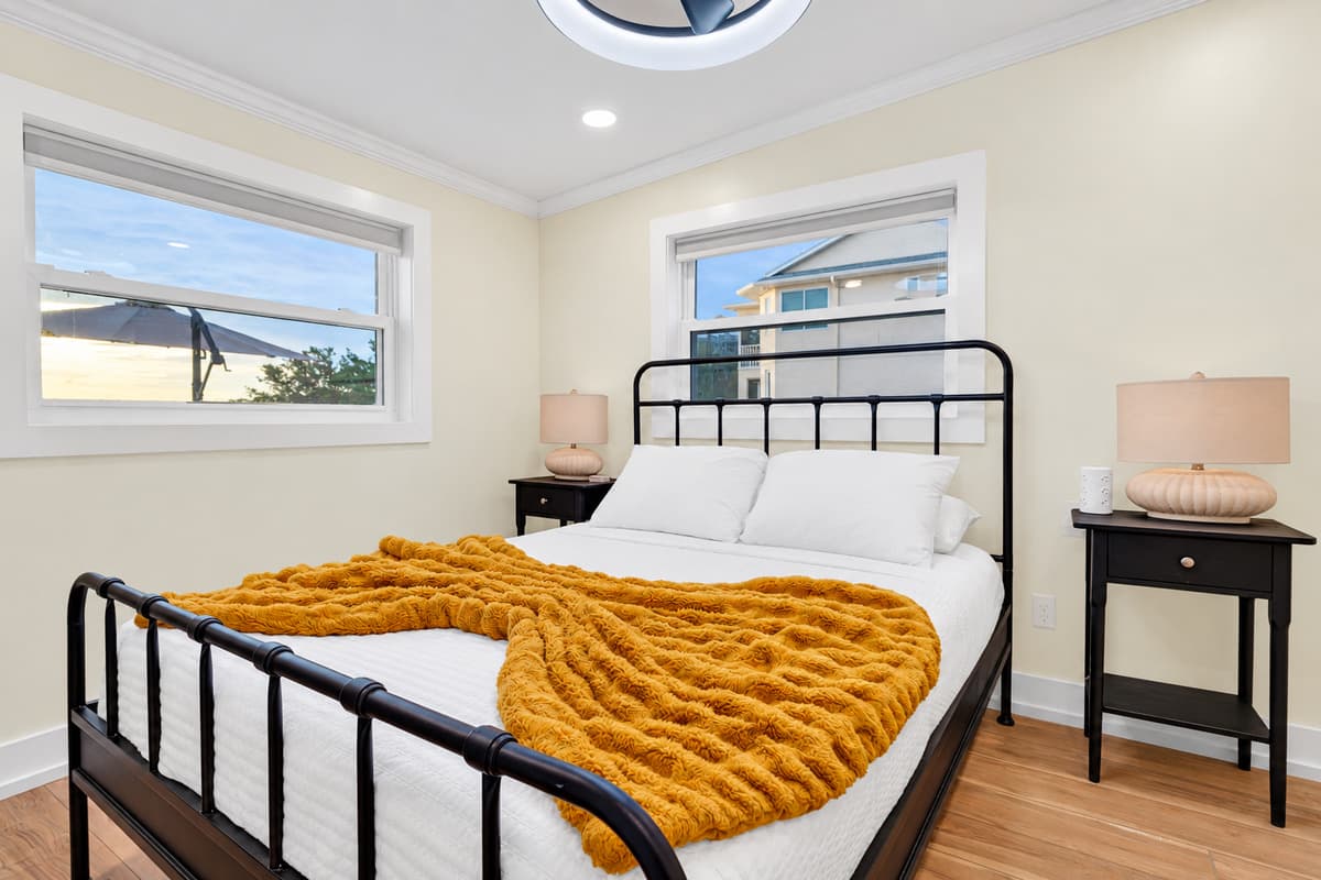 A black iron bed and soft textures. A queen bed with a black metal frame, a chunky knit throw and warm lamps. A cozy room with natural light from two windows.