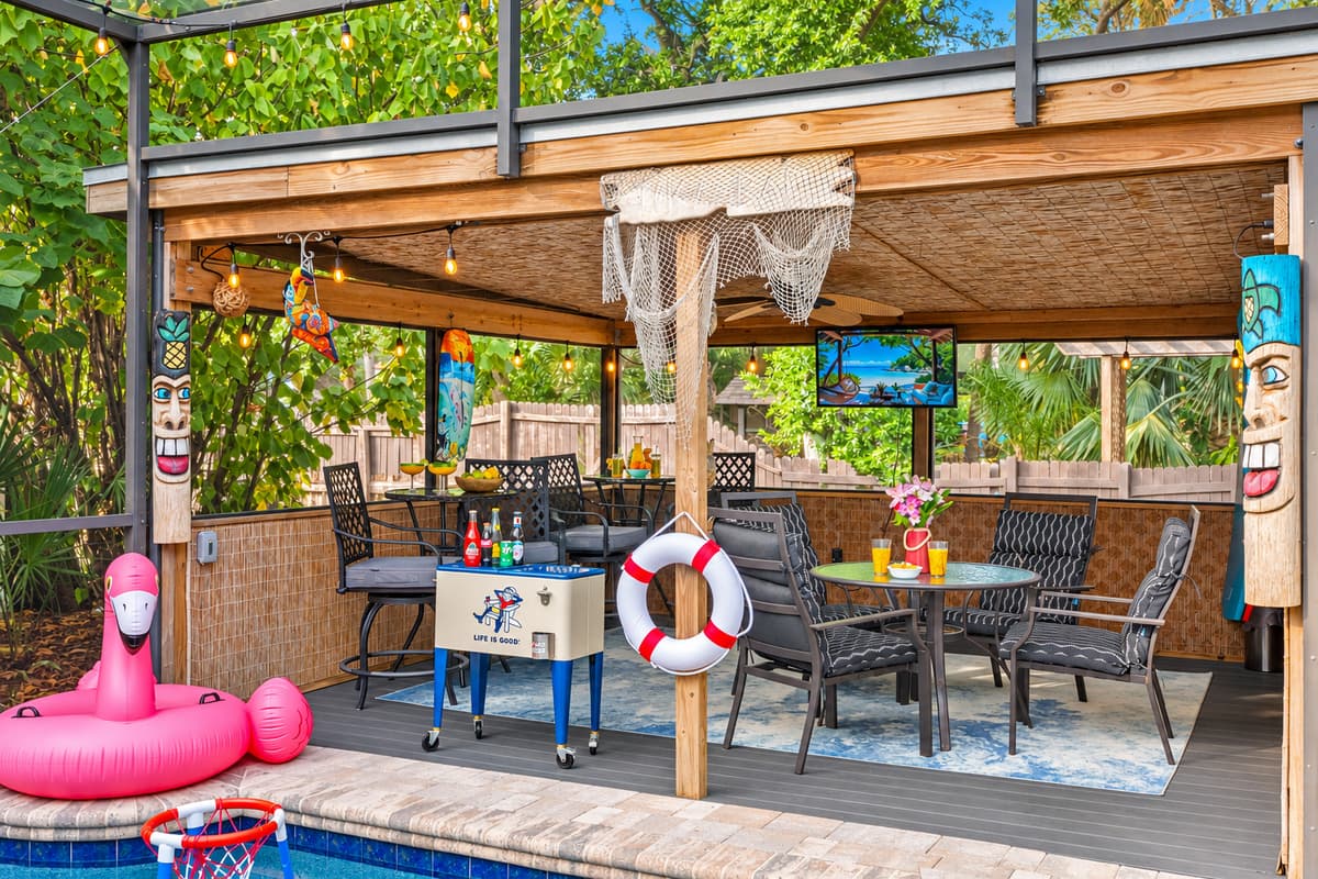 Poolside lounge area with tiki pillars, a beverage cooler, hanging décor, and a shaded dining setup under a bamboo-style ceiling.