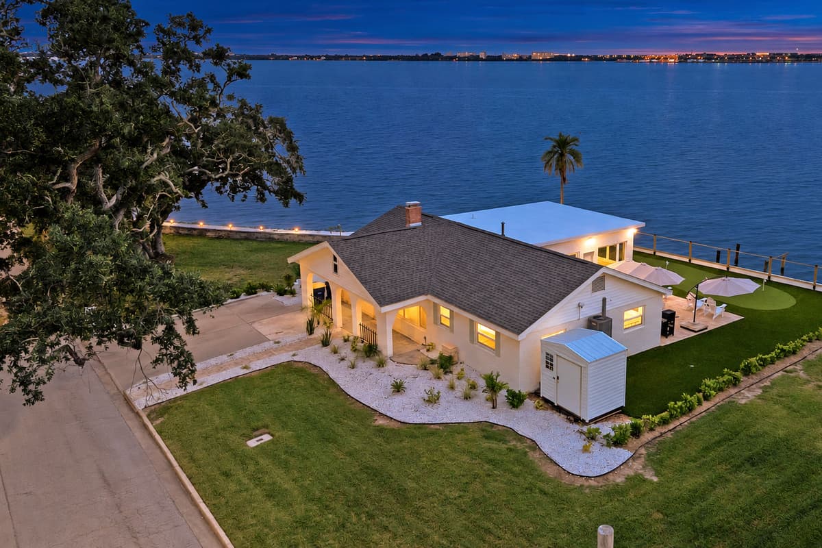Evening on the bay, lights twinkling. An aerial view at dusk shows the home lit up beside the water, with the putting green and patio glowing. Calm, private and unforgettable.