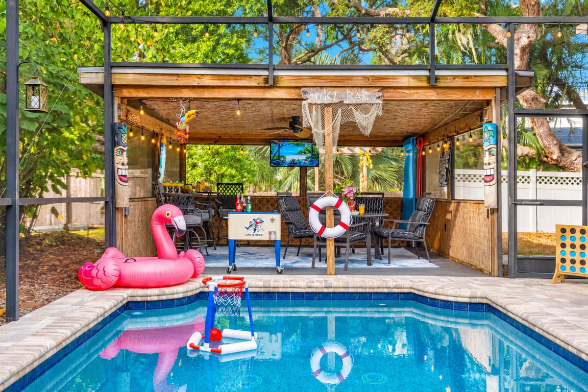 A fun, tiki-inspired patio complete with ample seating, bar area, TV, tropical décor, and a poolside view—perfect for relaxing or entertaining.