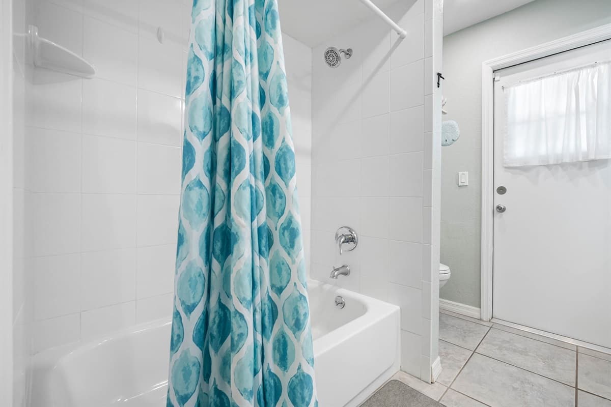 This full bathroom features a relaxing coastal design with dual vanities, granite countertops, and a beautifully tiled shower/tub combo accented by a vibrant blue curtain.