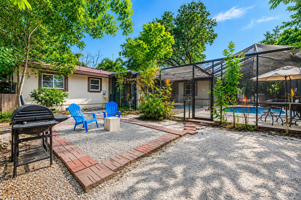 Wide view of the backyard featuring multiple outdoor gathering spaces, mature trees, and screened pool access.