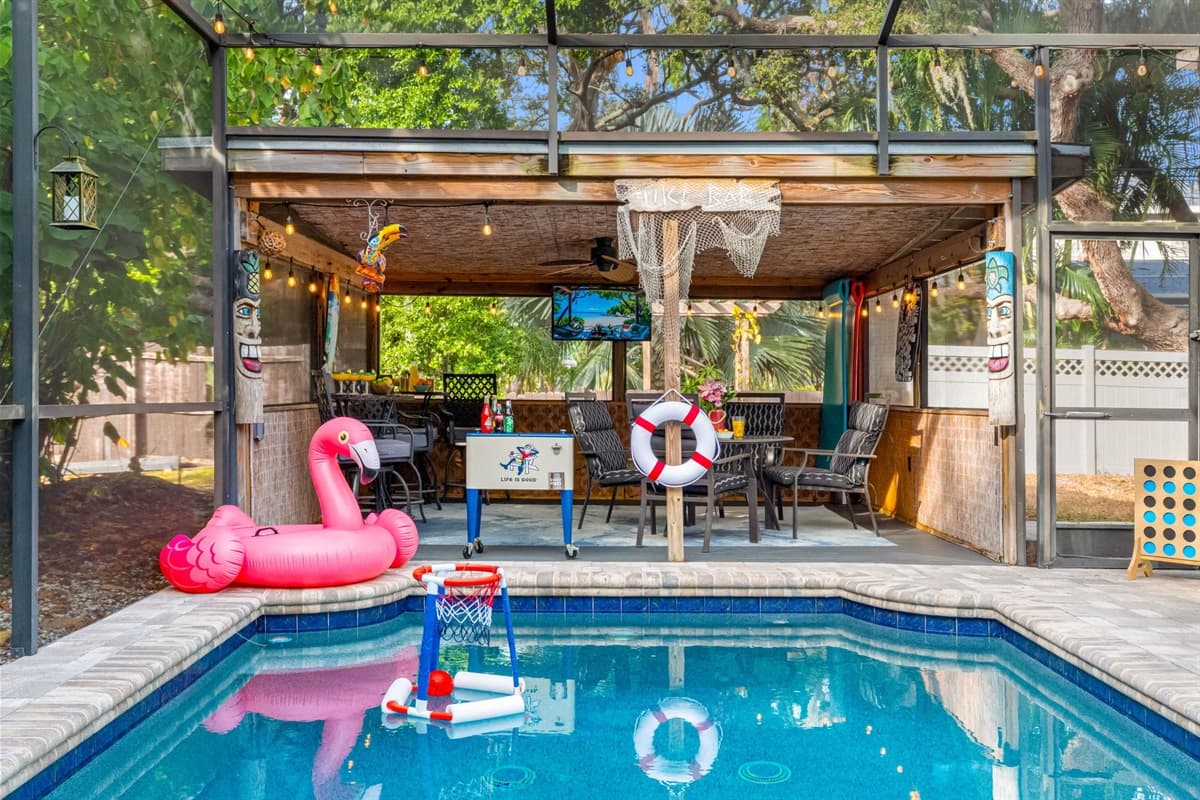 A fun, tiki-inspired patio complete with ample seating, bar area, TV, tropical décor, and a poolside view—perfect for relaxing or entertaining.
