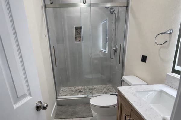 This beautifully updated bathroom features a sleek glass-enclosed shower with pebble tile accents, a modern wood vanity with a quartz countertop, and elegant brushed nickel fixtures.