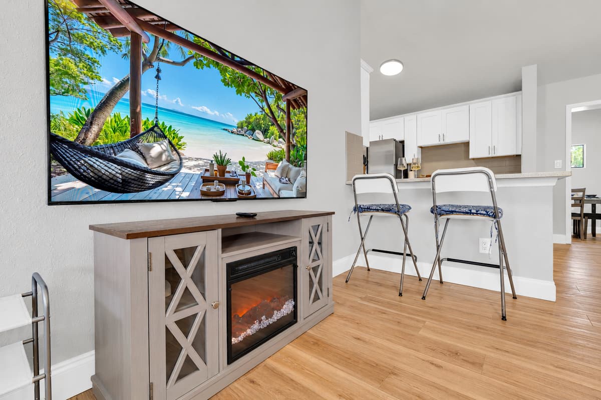 A mounted Smart TV over an electric fireplace, with counter stools pulled up beside it. Florida evenings rarely call for the fire, but it goes on anyway around December — mostly because somebody always turns it on.
