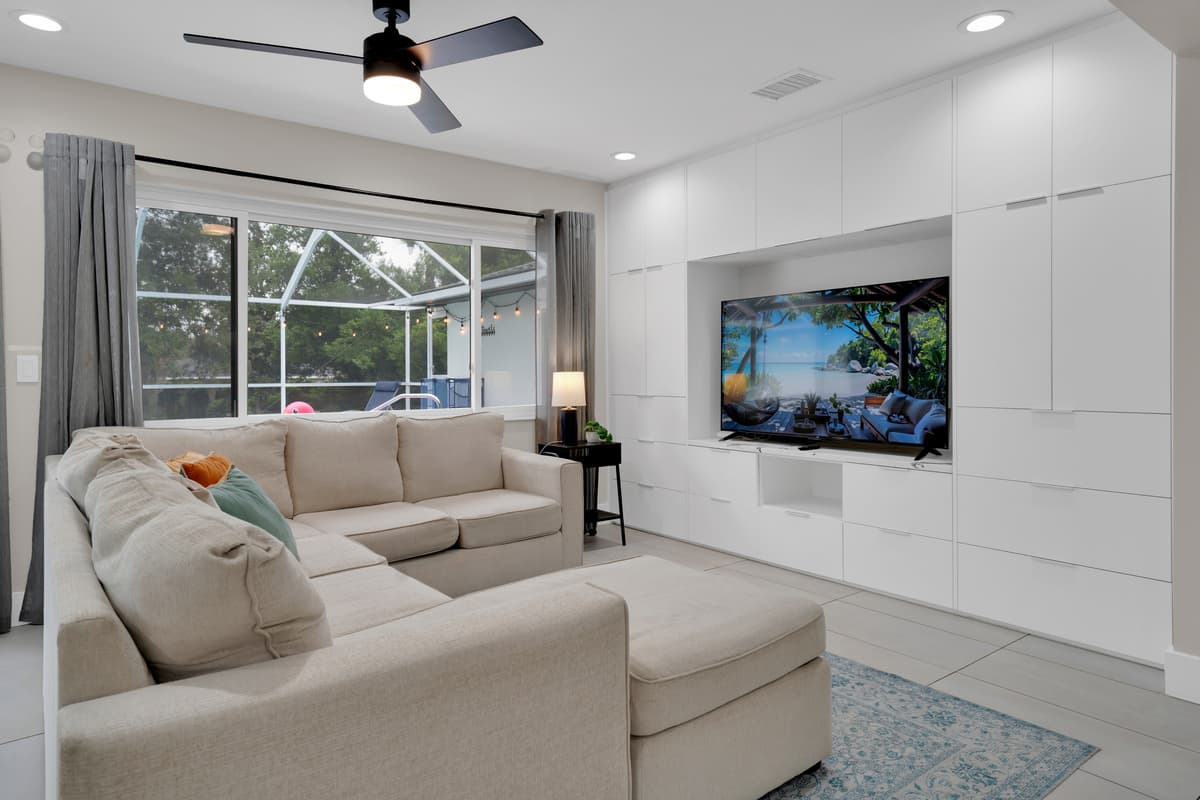 L-shaped sectional facing a wall-mounted TV, sun coming through floor-to-ceiling windows, a living room built around comfort, not just decoration.