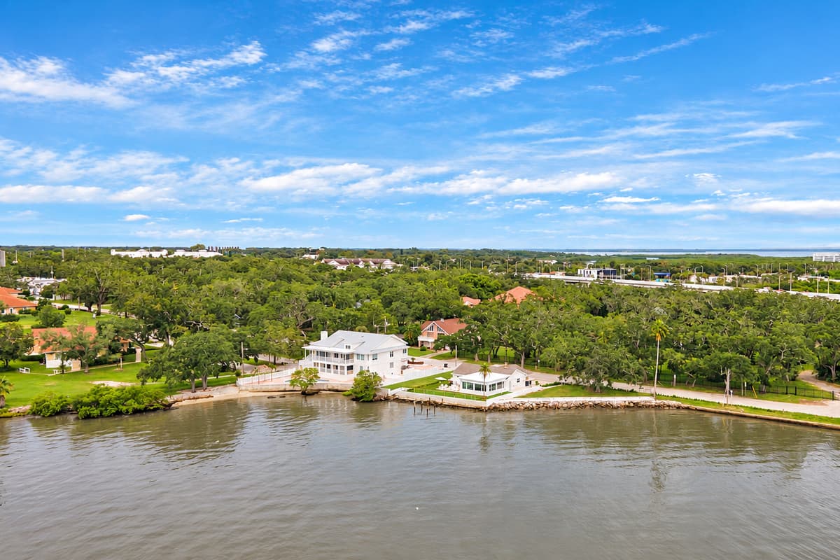 Tucked into the trees on Tampa Bay. A wide aerial view shows the home on the shoreline, surrounded by green trees and open water. Peaceful, residential and close to the bay.