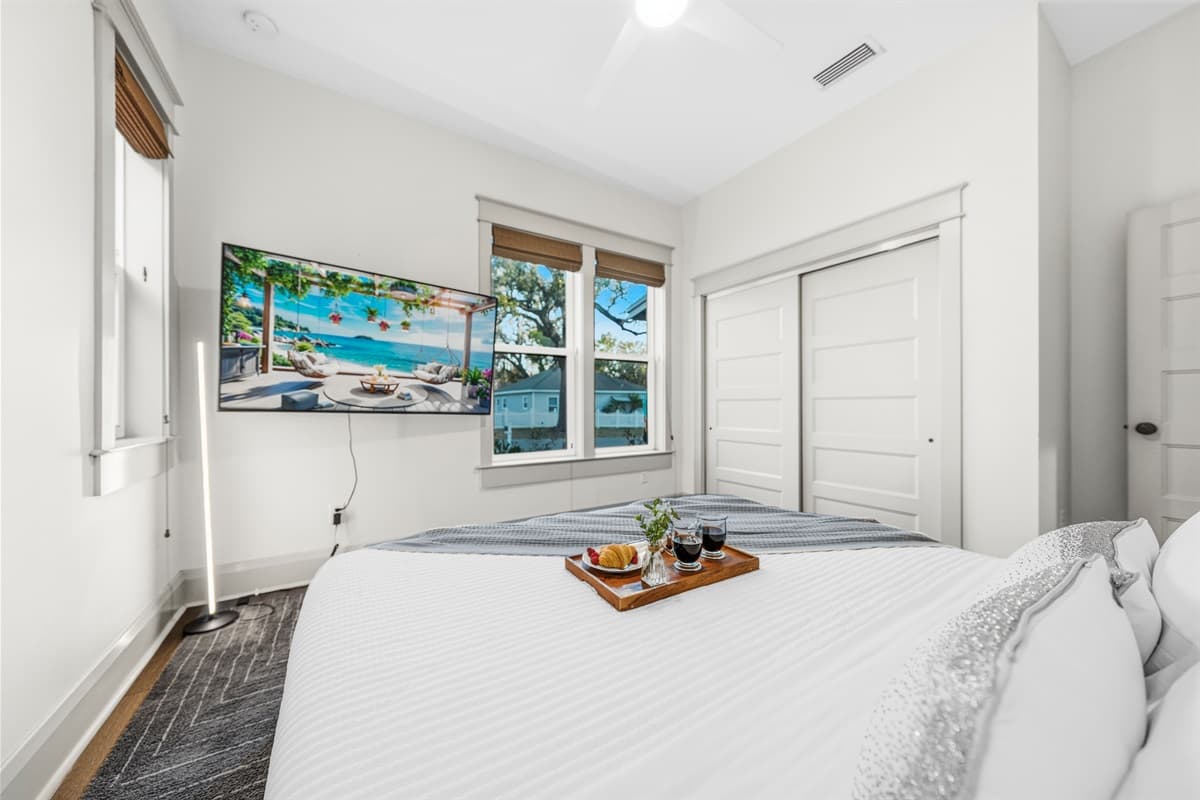 Bright white bedroom with a wall-mounted smart TV showing tropical paradise, a wooden breakfast tray, white shiplap accent wall, and bamboo shade. Brand-new vacation vibes in every inch of this Seminole Heights home.